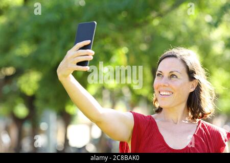 Felice donna adulta che prende selfie con smartphone in piedi nel parco in estate Foto Stock