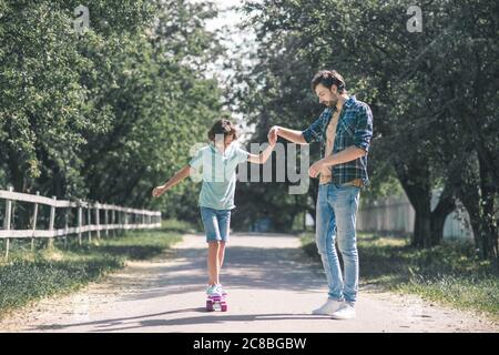 Teen che cavalcano uno skateboard, suo padre che tiene la mano e insegna Foto Stock