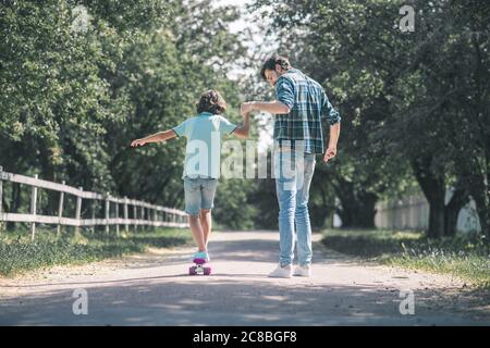 Ragazzo dai capelli scuri che cavalcano uno skateboard, suo padre lo tiene Foto Stock