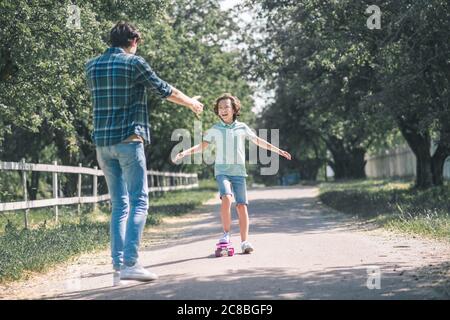 Ragazzo dai capelli scuri che cavalcano uno skateboard, suo padre si sente felice Foto Stock