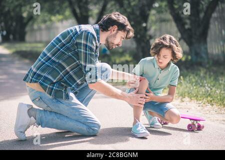 Uomo in una camicia a scacchi che esamina i suoi figli feriti ginocchio e guardando concentrato Foto Stock