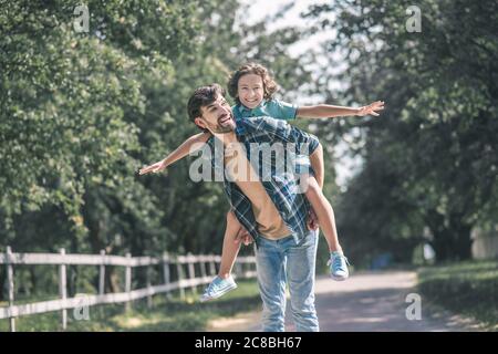 Ragazzo dai capelli scuri seduto sul suo padre indietro sentendosi come volare Foto Stock