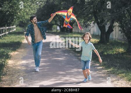 Ragazzo dai capelli scuri e suo padre che corrono dopo il colorato aquilone Foto Stock