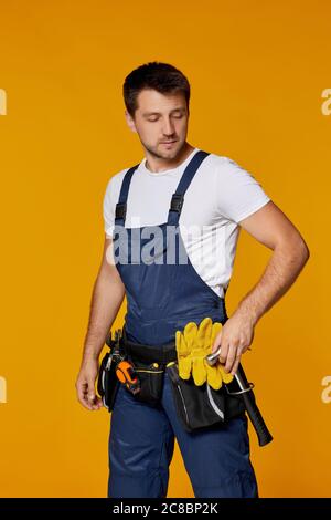 Giovane uomo di riparazione caucasico in elmetto giallo e martello di tenuta uniforme. Uomo a mano indossando cintura utensile isolato su sfondo arancione studio Foto Stock