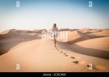 Bionda donna caucasica viaggiatore lasciando impronte in dune di sabbia quando camminare nel dessert in Oman Foto Stock