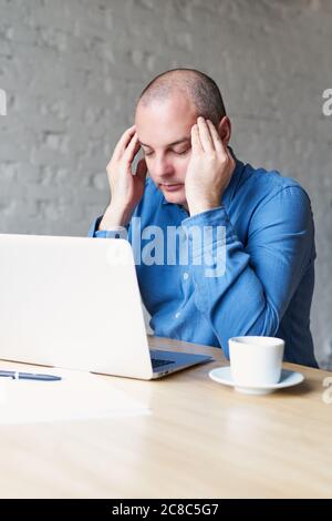 Un uomo soffre di mal di testa, si siede al lavoro in ufficio su un computer portatile, sfrega le mani con whiskey. Il concetto di lavoro eccessivo, stress, elaborazione Foto Stock