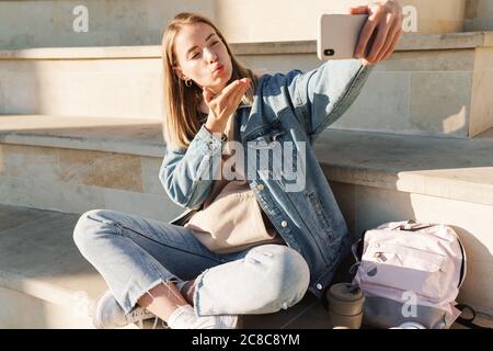 Allegra giovane ragazza bionda prendere un selfie con il telefono cellulare mentre si siede su passi otdoors, inviando bacio Foto Stock