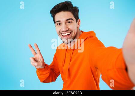 Immagine di felice giovane uomo setole isolato su sfondo blu parete prendere un selfie da fotocamera mostrando gesto di pace. Foto Stock