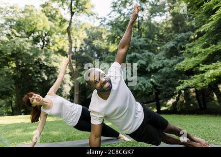 Giovane e donna gioiosa multietnica che si esercita in un parco, tenendo in posizione laterale con un braccio sollevato. Allenarsi in coppia all'aperto nel Foto Stock