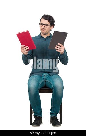 L'uomo pensivo studente, seduto su una sedia sembra confuso, scegliendo tra un libro tradizionale e un moderno tablet PC, isolato su bianco. Dilemma dell'istruzione Foto Stock