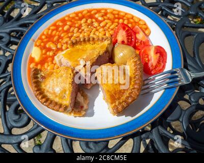 Pranzo snack un maiale Pie pomodori alla griglia fagioli e senape al forno su un piatto bianco con un bordo blu all'esterno su un tavolo di metallo Foto Stock