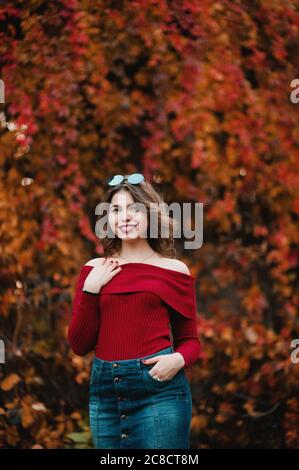 Bella giovane donna casual in occhiali che si trova tra giallo e rosso foglie nel parco pubblico, colori autunno sfondo Foto Stock