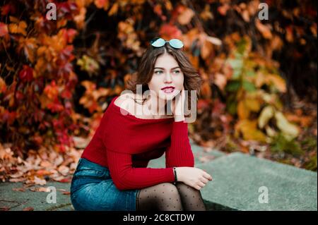 Bella giovane donna casual in occhiali che si trova tra giallo e rosso foglie nel parco pubblico, colori autunno sfondo Foto Stock