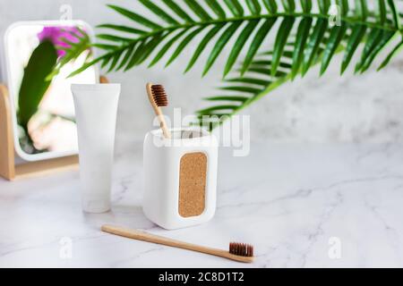 Spazzolini da denti in bambù in una tazza, dentifricio e specchio su un tavolo in marmo bianco in un bagno. Stile di vita sostenibile, concetto di igiene dentale. Foto Stock