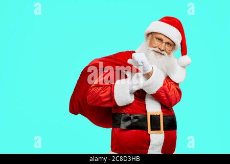 Vista frontale di Babbo Natale sorridente con borsa rossa e regali. Ritratto isolato di bel senior con barba bianca lunga in costume natalizio che posa su sfondo blu dello studio. Concetto di vacanze. Foto Stock