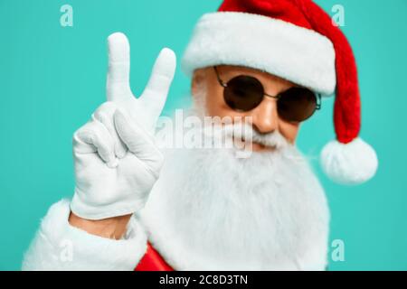 Vista frontale di Babbo Natale sorridente con la lunga barba bianca che mostra pace con due dita in su. Raccolto isolato di divertente uomo anziano in occhiali da sole che posano su sfondo blu studio. Concetto di vacanza, divertimento. Foto Stock