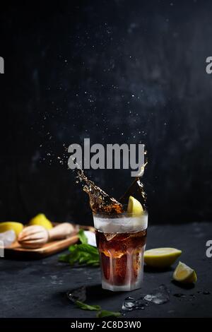 Cuba Libre o cocktail ghiacciato a lunga isola con bevande forti, cola, limone e ghiaccio in vetro, con spruzzi su sfondo scuro Foto Stock