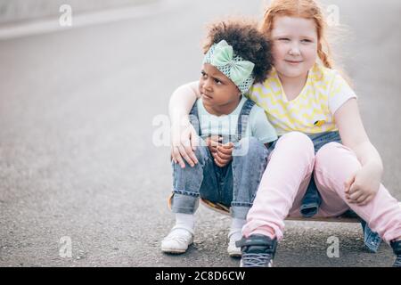 due amici che si siedono sullo skateboard all'aperto. Ragazza rossa e ragazza africana. Il concetto di amicizia e comunicazione interculturale Foto Stock