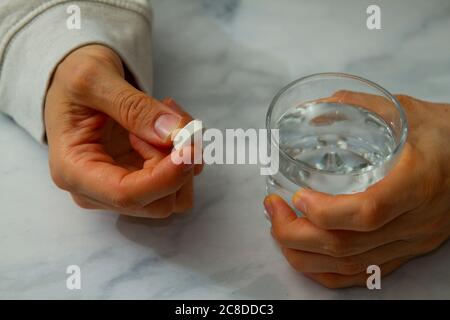 Una donna caucasica sta tenendo una pillola bianca in una mano e stringendo saldamente un bicchiere d'acqua dall'altra. Sta per prendere la pillola. Foto Stock