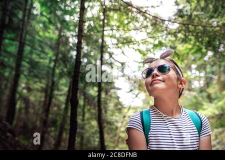 Carina bambina che indossa occhiali da sole e sorridente mentre si gode una giornata a piedi in una foresta in estate Foto Stock