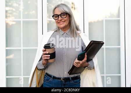 Sorridente donna d'affari anziana che tiene la tazza usa e getta con lima e intelligente telefono contro la porta dell'ufficio Foto Stock