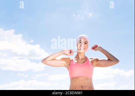 Donna sportiva e felice che indossa il copricapo mentre si alza in piedi contro il cielo blu Foto Stock