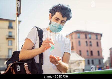 Uomo che indossa la maschera per il viso che lava le mani con l'igienizzatore mentre si è in piedi contro il cielo limpido Foto Stock