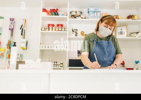 Donna panettiere indossando maschera di scrittura nel libro al banco cassa in negozio Foto Stock