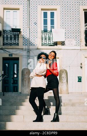 Amici femminili che indossano occhiali da sole in piedi sui gradini contro la costruzione in città Foto Stock