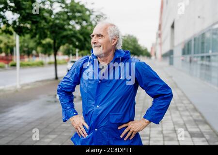 Uomo anziano che indossa un impermeabile blu che guarda lontano mentre si sta in piedi sentiero in città Foto Stock