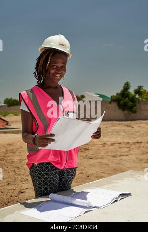 Sorridente architetto femminile che tiene il modello mentre si sta in piedi contro il cielo chiaro in loco durante la giornata di sole Foto Stock