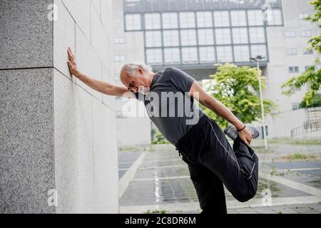 Uomo anziano che allunga la gamba mentre si sta a parete sul sentiero in città Foto Stock