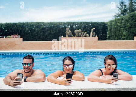 Amici durante una festa in piscina utilizzando gli smartphone Foto Stock