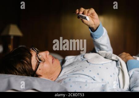 Donna anziana che guarda il termometro mentre si sdraiava sul letto a. casa Foto Stock