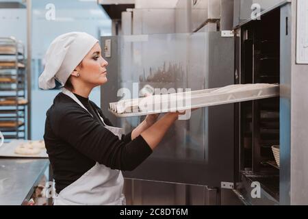 Panettiere femmina che inserisce la teglia nel forno della panetteria Foto Stock