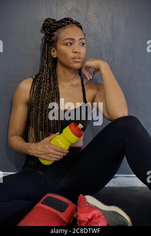 Atleta femminile premurosa con biberon intrecciato per la presa dei capelli mentre si è seduti contro il muro in palestra Foto Stock