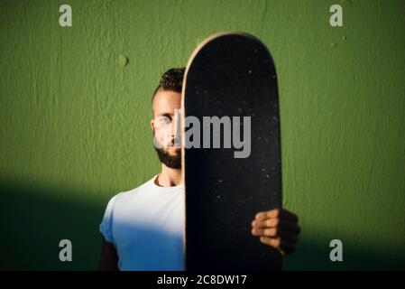 Bel giovane uomo che tiene lo skateboard mentre si trova contro il muro verde Foto Stock