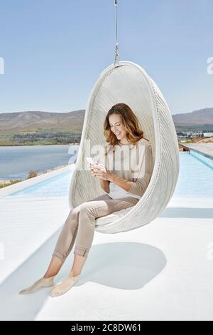 Donna sorridente seduta in sedia pensile sopra la piscina utilizzando smartphone Foto Stock