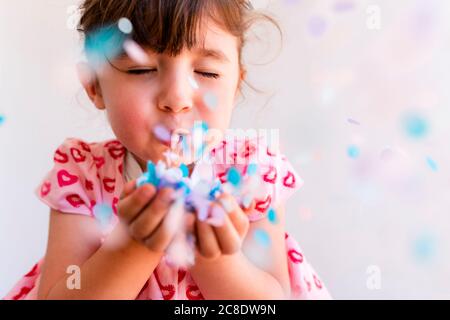 Primo piano di ragazza carina con gli occhi chiusi soffiando confetti da mani su sfondo bianco Foto Stock