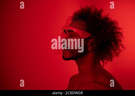 Giovane uomo con gli occhi chiusi indossando una maschera a LED rossa contro il muro a casa Foto Stock