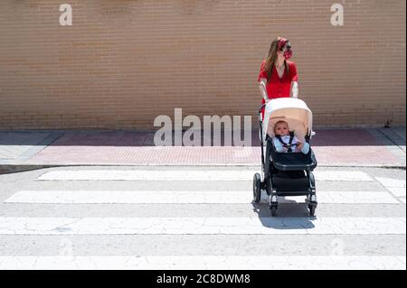 La madre indossa una maschera che spinge il figlio nel carrello del bambino mentre cammina sulla strada Foto Stock