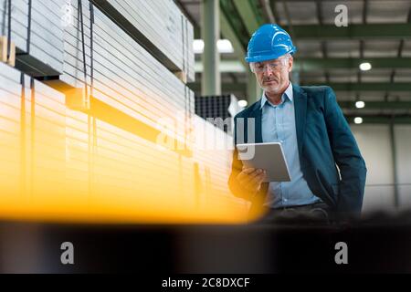 Uomo d'affari maturo che indossa casco e occhiali di sicurezza in un in fabbrica utilizzando un tablet Foto Stock