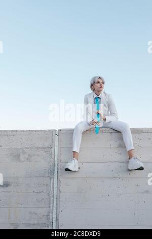 Donna giovane premurosa che indossa un vestito bianco seduto su una parete di ritegno contro il cielo limpido Foto Stock