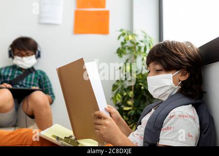 Ragazzi che indossano maschere di studio mentre si siedono a distanza a scuola Foto Stock