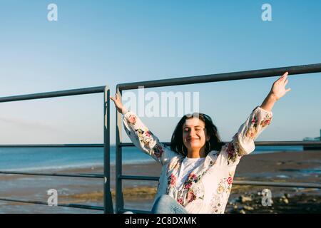 Ragazza adolescente sorridente con le braccia sollevate seduta da ringhiera contro mare Foto Stock