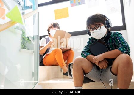 Ragazzi che indossano maschere seduti a distanza sui gradini della scuola Foto Stock