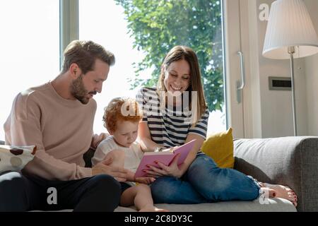 Donna sorridente che legge libro dell'immagine mentre sedendo dal figlio e. uomo sul divano nel soggiorno di casa Foto Stock