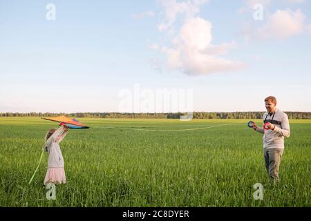 Padre che tiene rocchetti mentre figlia vola aquiloni sul paesaggio verde Foto Stock