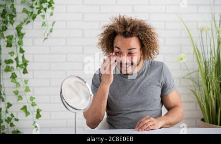 Sorridente giovane uomo che applica le macchie dell'occhio sul viso mentre si siede a casa Foto Stock