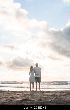 Coppia giovane amorevole con le braccia intorno a guardare il mare contro cielo nuvoloso Foto Stock